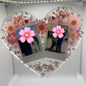 May include: A heart-shaped resin picture frame with two Polaroid photos, pink flowers, and silver glitter. The frame has a silver chain for hanging and gold charms with the letters "A" and "C".