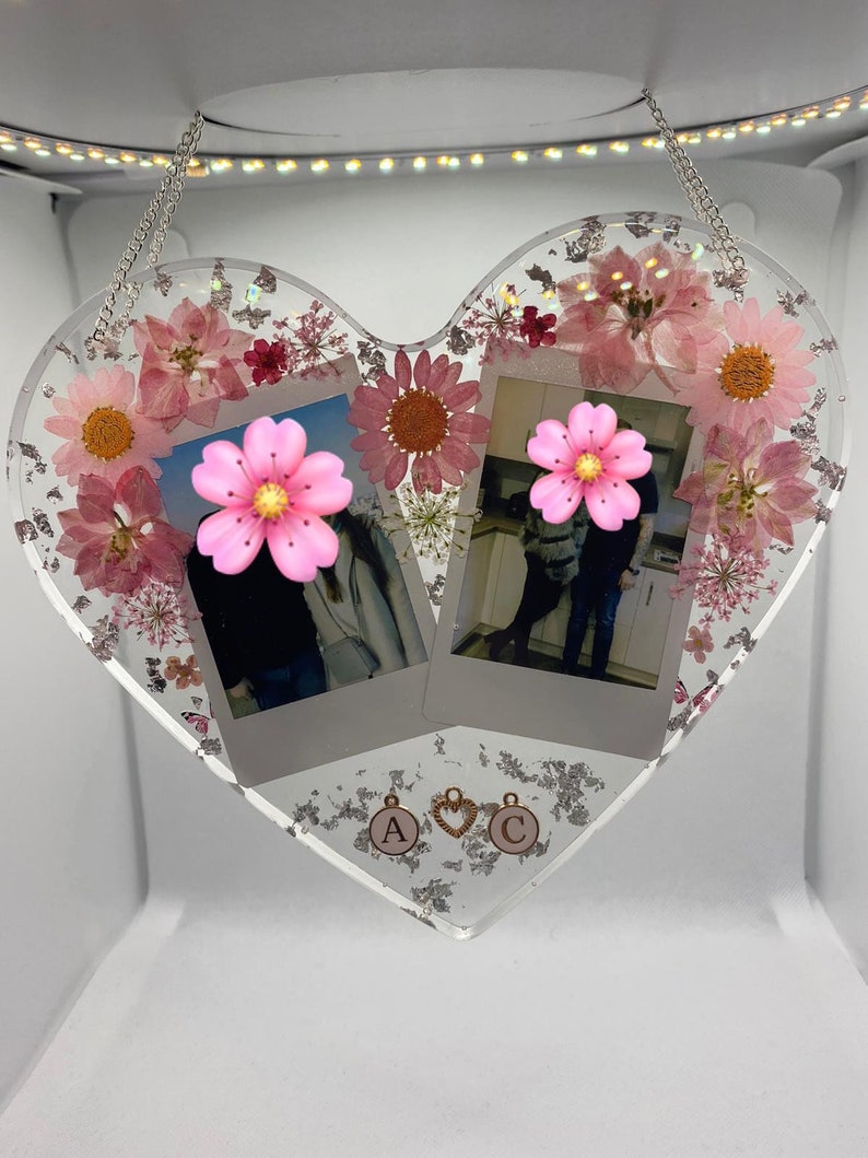 May include: A heart-shaped resin picture frame with two Polaroid photos, pink flowers, and silver glitter. The frame has a silver chain for hanging and gold charms with the letters "A" and "C".