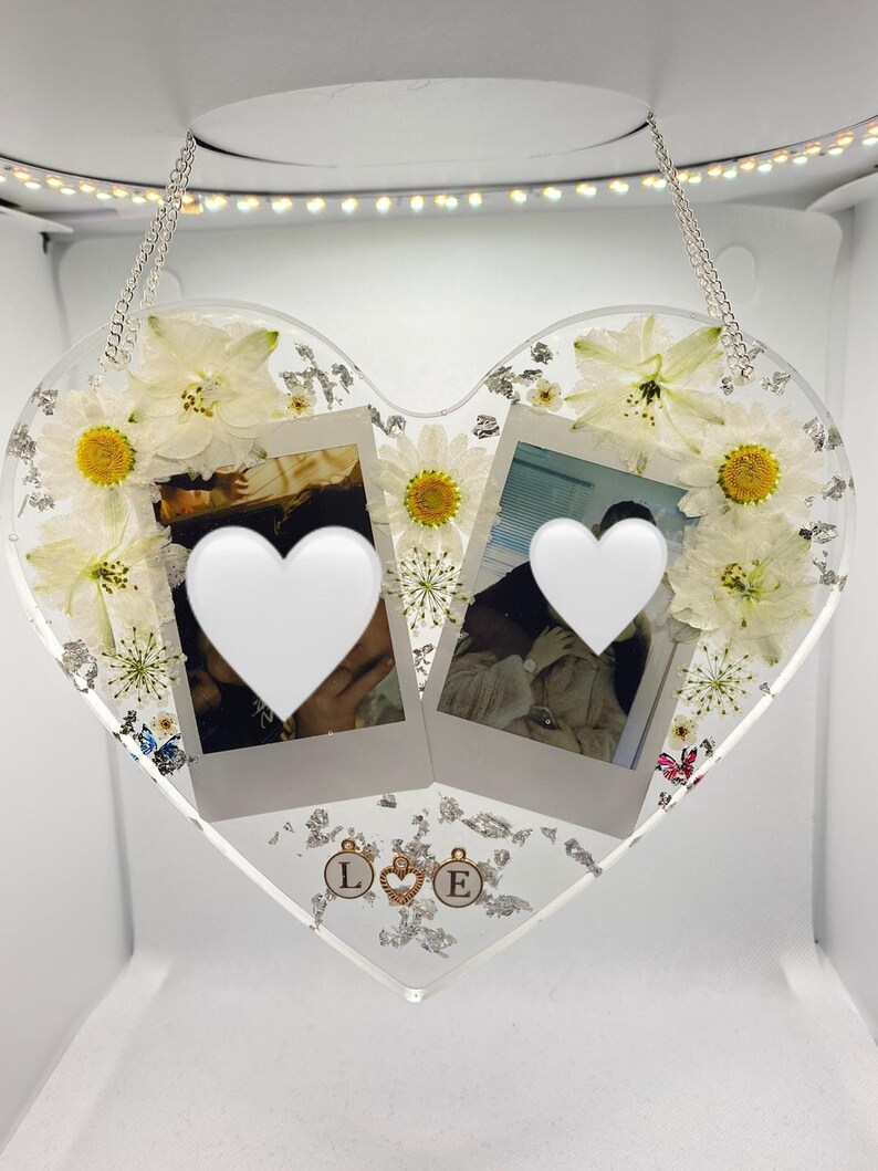 May include: A white heart-shaped resin ornament with two Polaroid photos, white flowers, silver glitter, and gold charms with the letters "L" and "E".