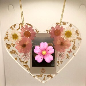 May include: A heart-shaped resin ornament with a gold chain, featuring pressed pink and white flowers, gold leaf accents, and a small photo inside.
