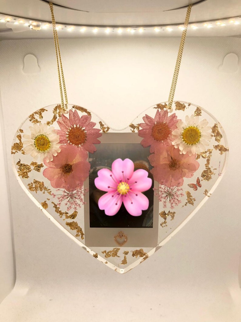 May include: A heart-shaped resin ornament with a gold chain, featuring pressed pink and white flowers, gold leaf accents, and a small photo inside.