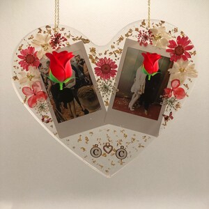 May include: A heart-shaped resin ornament with two framed photos, dried flowers, and gold leaf accents. The photos feature a couple and a red rose. The ornament has two charms with the letter "C" and a heart.