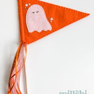 May include: Orange felt pennant with a pink ghost and silver glitter stars. The pennant is attached to a wooden stick with orange and pink ribbon streamers.