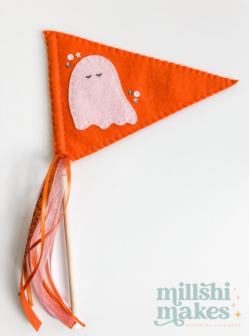 May include: Orange felt pennant with a pink ghost and silver glitter stars. The pennant is attached to a wooden stick with orange and pink ribbon streamers.