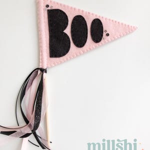 May include: Pink felt pennant with the word "BOO" in black felt letters. The pennant has a wooden stick and pink and black ribbon streamers.