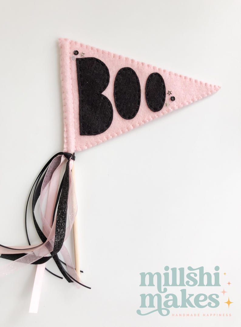 May include: Pink felt pennant with the word "BOO" in black felt letters. The pennant has a wooden stick and pink and black ribbon streamers.