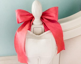 May include: A white ceramic light switch cover with a pink bow tied around it. The switch cover is shaped like a tooth and is attached to a white wall.