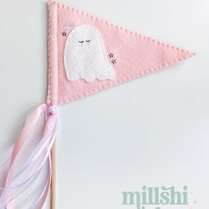 May include: A pink felt pennant with a white ghost applique and black embroidered eyes. The pennant has a pink and white ribbon attached to a wooden stick.