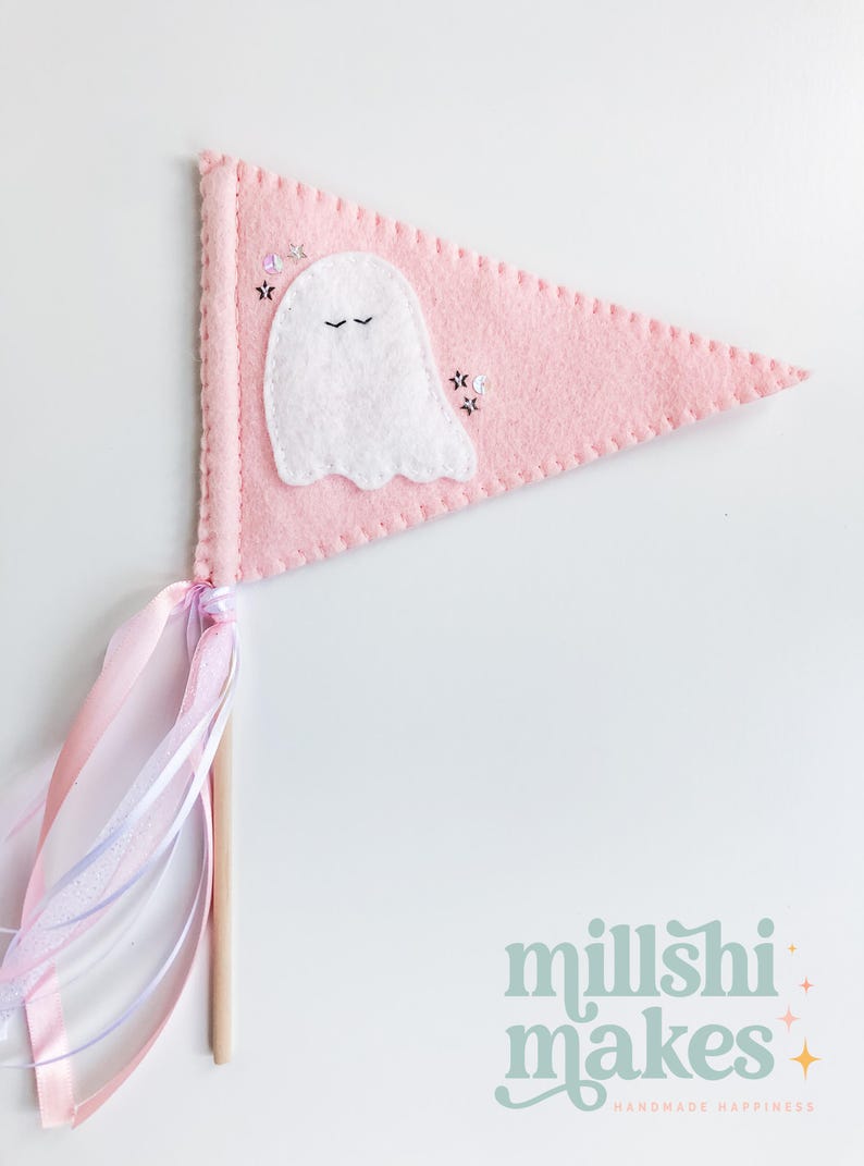 May include: A pink felt pennant with a white ghost applique and black embroidered eyes. The pennant has a pink and white ribbon attached to a wooden stick.