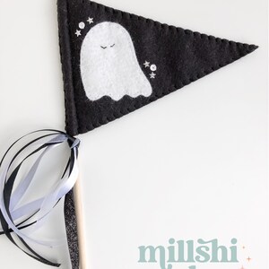 May include: A black felt pennant with a white ghost and silver glitter stars. The pennant is attached to a wooden stick with black and white ribbon streamers. The text "millshi makes" is printed in light blue with pink and yellow stars.