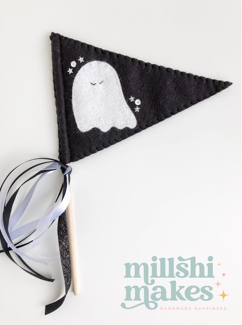 May include: A black felt pennant with a white ghost and silver glitter stars. The pennant is attached to a wooden stick with black and white ribbon streamers. The text "millshi makes" is printed in light blue with pink and yellow stars.