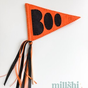 May include: Orange felt pennant with the word "BOO" in black felt letters. The pennant is attached to a wooden stick with orange, black, and white ribbon streamers.