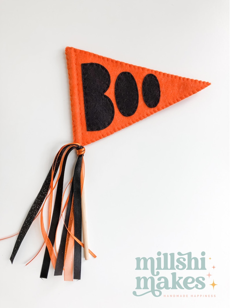 May include: Orange felt pennant with the word "BOO" in black felt letters. The pennant is attached to a wooden stick with orange, black, and white ribbon streamers.