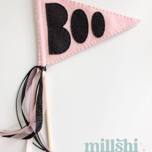 May include: Pink felt pennant with black felt letters spelling "BOO" on a white background. The pennant is attached to a wooden stick with pink and black ribbon streamers.