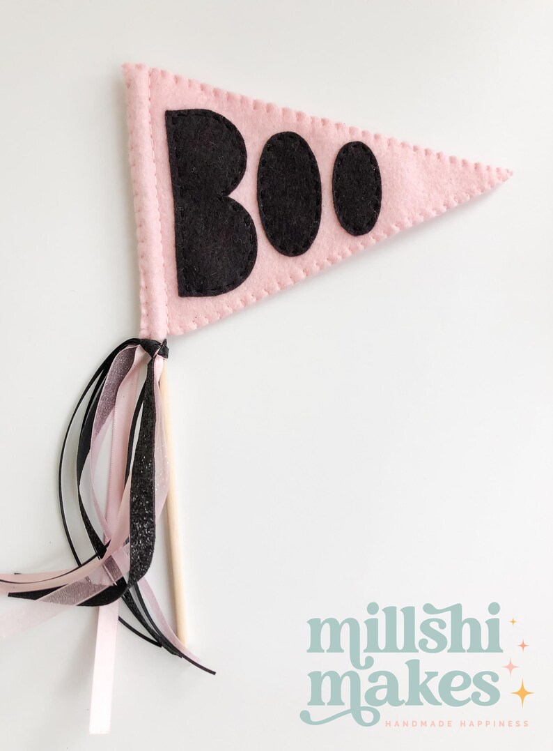 May include: Pink felt pennant with black felt letters spelling "BOO" on a white background. The pennant is attached to a wooden stick with pink and black ribbon streamers.