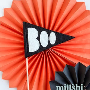 May include: A black felt pennant with the word "BOO" in white felt letters. The pennant is attached to a wooden stick and is displayed against a bright orange paper fan.