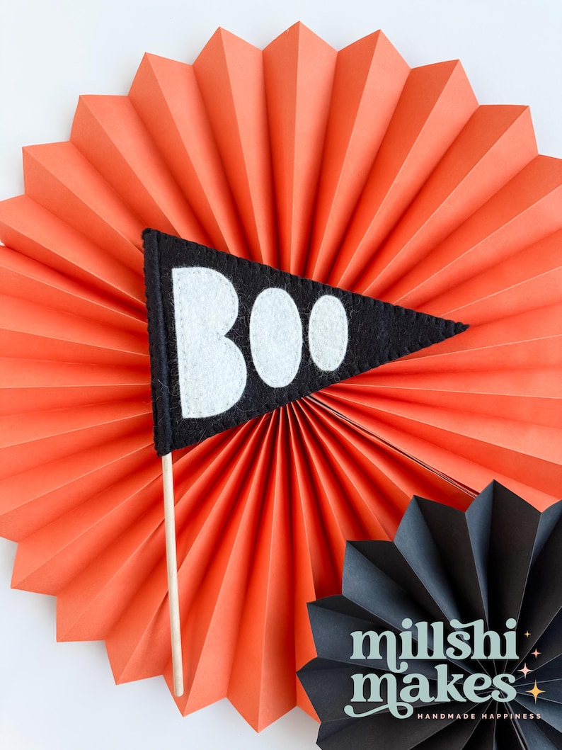 May include: A black felt pennant with the word "BOO" in white felt letters. The pennant is attached to a wooden stick and is displayed against a bright orange paper fan.