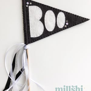 May include: Black felt pennant with white felt letters spelling "BOO" and silver glitter stars. The pennant is attached to a wooden stick with black, white, and silver ribbon streamers.
