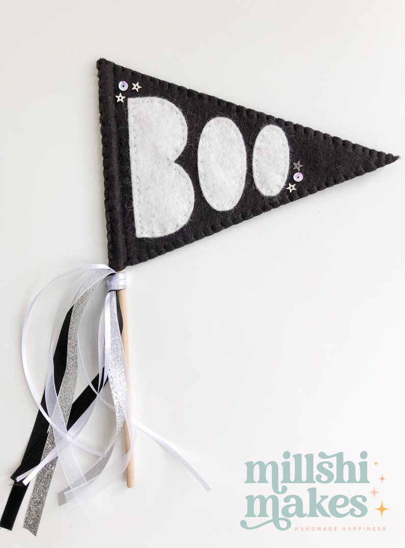 May include: Black felt pennant with white felt letters spelling "BOO" and silver glitter stars. The pennant is attached to a wooden stick with black, white, and silver ribbon streamers.