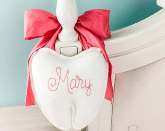 May include: A white plush tooth-shaped pillow featuring the name Mary embroidered in pink thread. The pillow is tied to a white bedpost with a large, vibrant pink fabric bow. The text millshi makes is visible in the lower right corner.