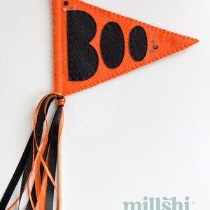 May include: Orange felt pennant with the word "BOO" in black felt letters and glitter accents. The pennant is attached to a wooden stick with orange, black, and white ribbon streamers.