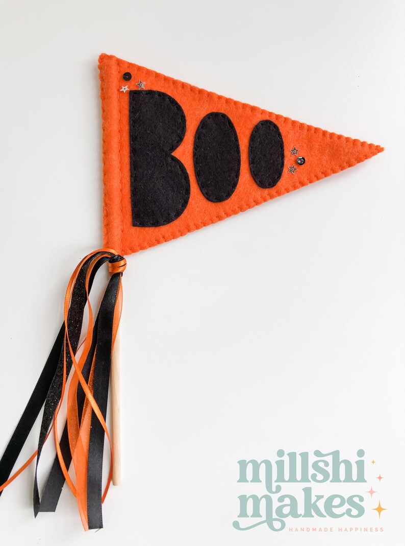 May include: Orange felt pennant with the word "BOO" in black felt letters and glitter accents. The pennant is attached to a wooden stick with orange, black, and white ribbon streamers.