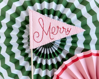 May include: A pink felt pennant with the word "Merry" embroidered in red thread. The pennant is attached to a wooden stick and is sitting on top of a green and white chevron patterned paper fan.