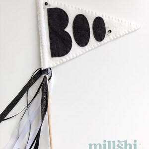 May include: White felt pennant with black felt letters spelling "BOO" and black glitter accents. The pennant is attached to a wooden stick with black and white ribbon streamers.