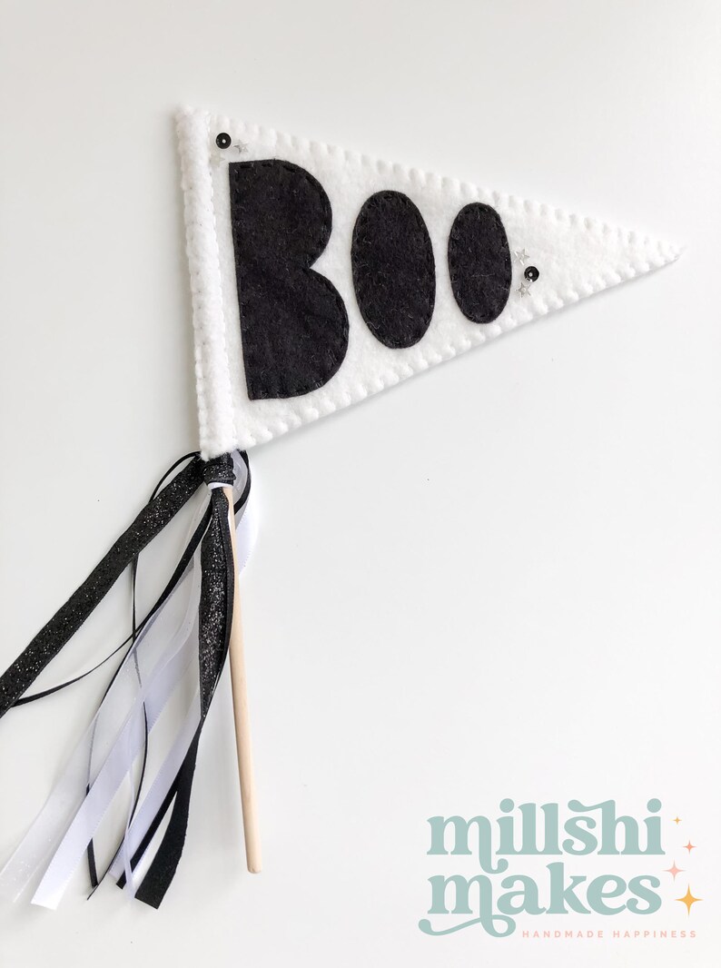 May include: White felt pennant with black felt letters spelling "BOO" and black glitter accents. The pennant is attached to a wooden stick with black and white ribbon streamers.