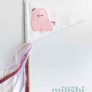 May include: A white felt pennant with a pink ghost and pink flowers embroidered on it. The pennant is attached to a wooden stick with pink and white ribbons.