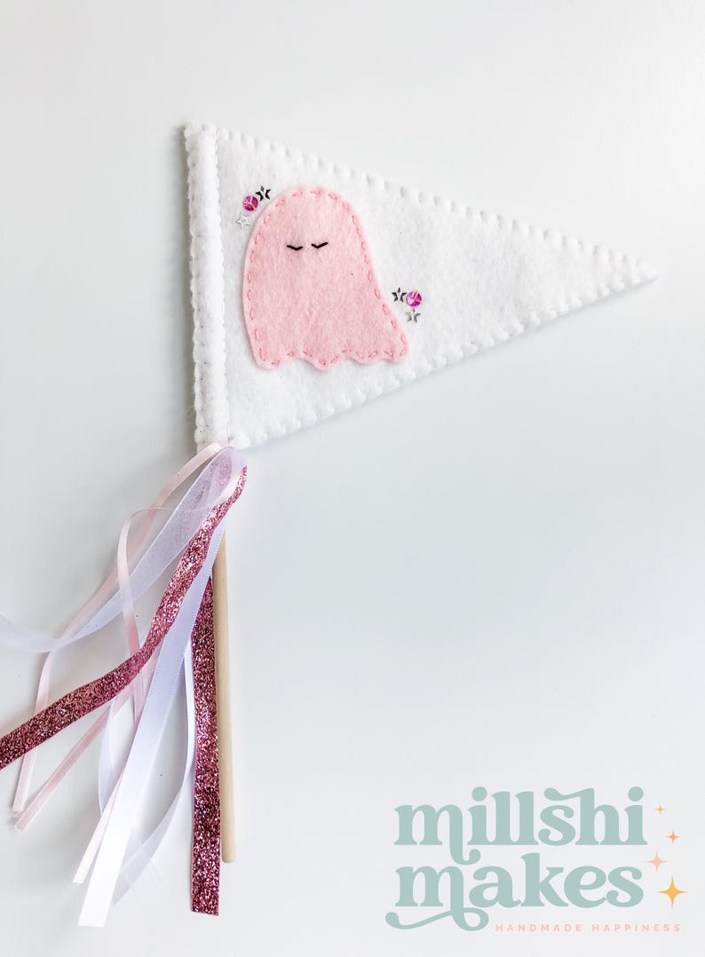 May include: A white felt pennant with a pink ghost and pink flowers embroidered on it. The pennant is attached to a wooden stick with pink and white ribbons.
