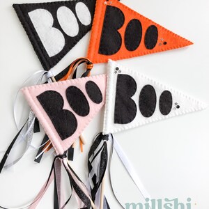 May include: Three felt pennant flags with the word "BOO" in black letters on a white, pink, and orange background. Each flag is attached to a wooden stick with black, white, and pink ribbon streamers.
