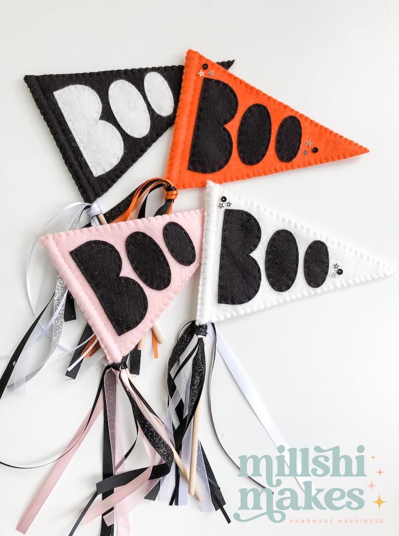 May include: Three felt pennant flags with the word "BOO" in black letters on a white, pink, and orange background. Each flag is attached to a wooden stick with black, white, and pink ribbon streamers.