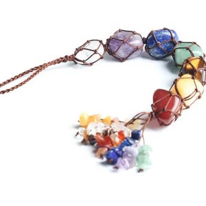 Könnte beinhalten: Eine sieben-Chakra-Kristallkette mit einer braunen Schnur und einem Quasten aus kleineren Kristallen. Die Kristalle sind in braune Schnur gewickelt und in einem Regenbogenmuster angeordnet.