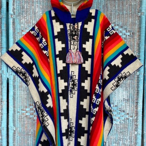 Könnte beinhalten: Ein farbenfrohes Poncho mit Kapuze, mit einem geometrischen Muster in Schwarz, Weiß, Rot, Blau und Gelb. Der Poncho hat einen Fransenbesatz und Quasten-Details, ideal für einen Bohemian- oder Südwest-Stil.
