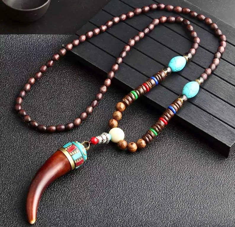 K&ouml;nnte beinhalten: Eine lange Holzperlenkette mit einem braunen Holz-Zahn-Anh&auml;nger. Die Kette hat bunte Perlen und t&uuml;rkisfarbene Akzente.
