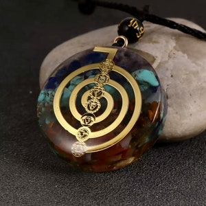 K&ouml;nnte beinhalten: Ein goldfarbener Anh&auml;nger mit einem Sieben-Chakra-Symbol, einer durchsichtigen Harzbasis, die mit verschiedenen farbigen Steinen gef&uuml;llt ist. Der Anh&auml;nger ist an einer schwarzen Schnur mit einer schwarzen Perle.