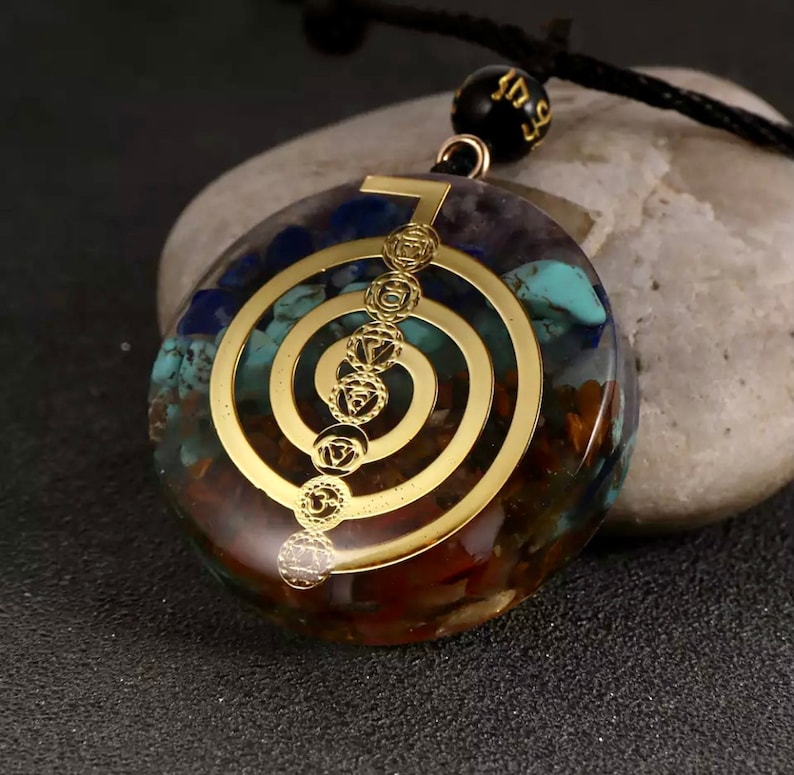 K&ouml;nnte beinhalten: Ein goldfarbener Anh&auml;nger mit einem Sieben-Chakra-Symbol, einer durchsichtigen Harzbasis, die mit verschiedenen farbigen Steinen gef&uuml;llt ist. Der Anh&auml;nger ist an einer schwarzen Schnur mit einer schwarzen Perle.