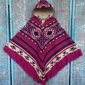 Könnte beinhalten: Ein Kapuzenponcho in Magenta-, Lila- und Cremetönen mit geometrischen Mustern und Fransen. Der Poncho hat einen Knopfverschluss und eine gemusterte Kapuze. Das Design umfasst Streifen und Rauten.