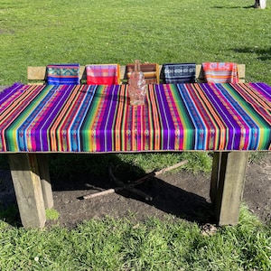 Op de afbeelding: Een kleurrijk gestreept tafelkleed met franjes aan de randen, gelegd op een houten picknicktafel. Het tafelkleed is paars, oranje, geel, groen en blauw. Er liggen vijf kleurrijke servetten gevouwen op de tafel.