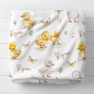 May include: White baby blanket with a repeating pattern of yellow ducklings, butterflies, and green leaves. The name "Ryan" is written in brown script across the blanket. The blanket has a soft, plush texture.