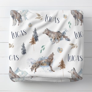 May include: White baby blanket with a watercolor design featuring wolves, trees, and floral accents. The name "LUCAS" is repeated across the blanket in a stylish font. The blanket has a soft, cozy appearance.