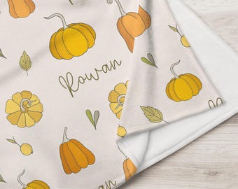 May include: A soft, folded blanket featuring a repeating pattern of yellow and orange pumpkins and green leaves on a light cream background. The name Rowan is printed in a dark brown cursive script among the pumpkin illustrations.