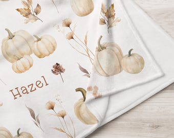 May include: A white fleece blanket featuring a repeating watercolor pattern of beige pumpkins and brown autumn leaves. The name Hazel is printed in a brown serif font on the fabric. The blanket is folded to show the soft texture and underside.