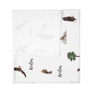 May include: White baby blanket with a repeating pattern of eagles, mountains, trees, and the name "Arden" written in black.