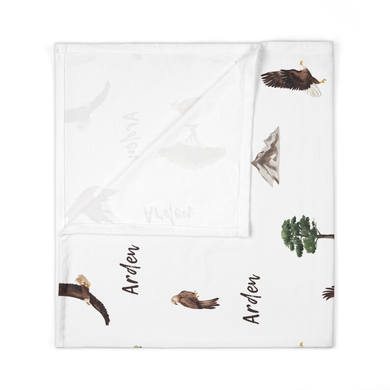 May include: White baby blanket with a repeating pattern of eagles, mountains, trees, and the name "Arden" written in black.