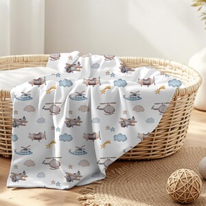 May include: A white baby blanket with a repeating pattern of airplanes, helicopters, clouds, stars, and shooting stars in shades of brown, blue, and yellow. The blanket is draped over a wicker basket.