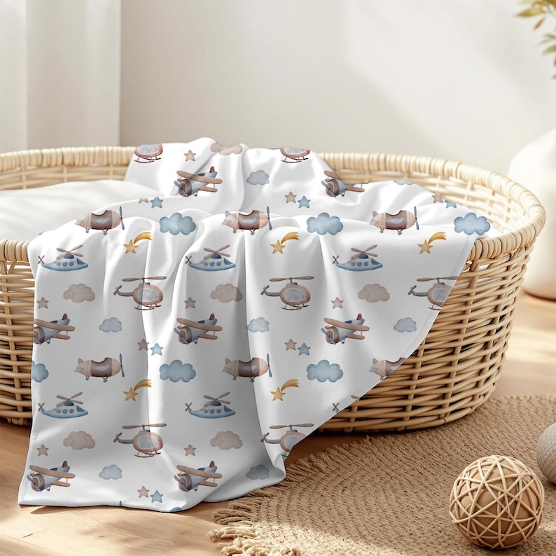 May include: A white baby blanket with a repeating pattern of airplanes, helicopters, clouds, stars, and shooting stars in shades of brown, blue, and yellow. The blanket is draped over a wicker basket.
