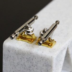 May include: Two gold-coloured fingerboard trucks with silver axles and white bushings. The trucks are on a white surface with black speckles.