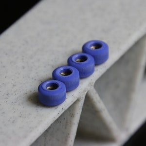Puede incluir: Cuatro ruedas de fingerboard azules alineadas en una superficie blanca. Las ruedas son de plástico duro y tienen un acabado liso.